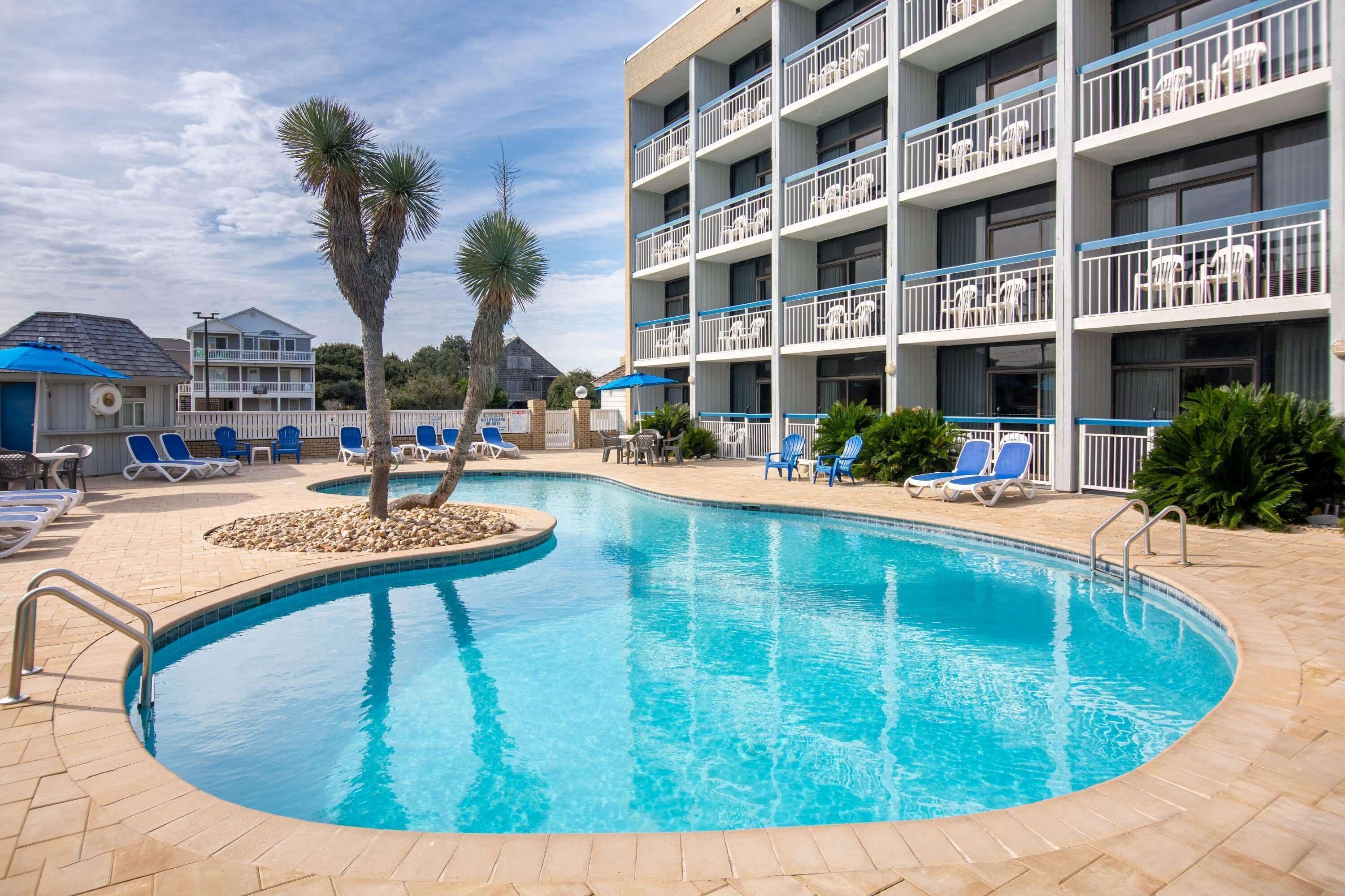 A swimming pool with lounge chairs and palm trees, surrounded by a multi-story building with balconies.