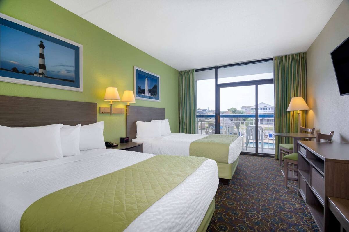 Habitación de hotel con dos camas, un escritorio, una silla, un televisor y un balcón con vistas a edificios y una masa de agua.