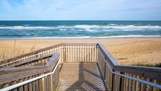Een houten looppad dat naar een zandstrand leidt, met de oceaan op de achtergrond.