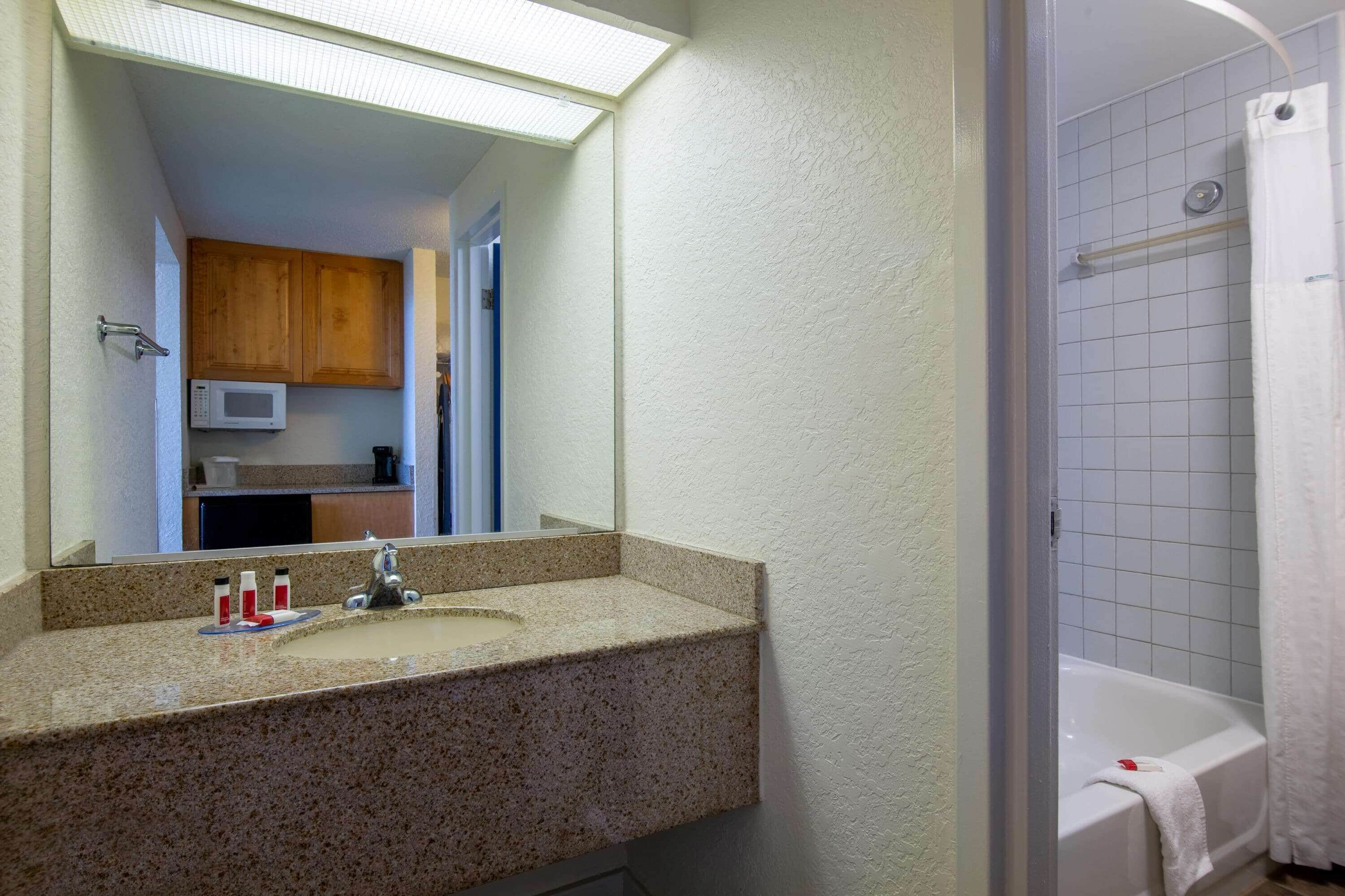 Baño con lavamanos de encimera de granito, espejo, ducha con cortina blanca y microondas sobre el lavamanos.