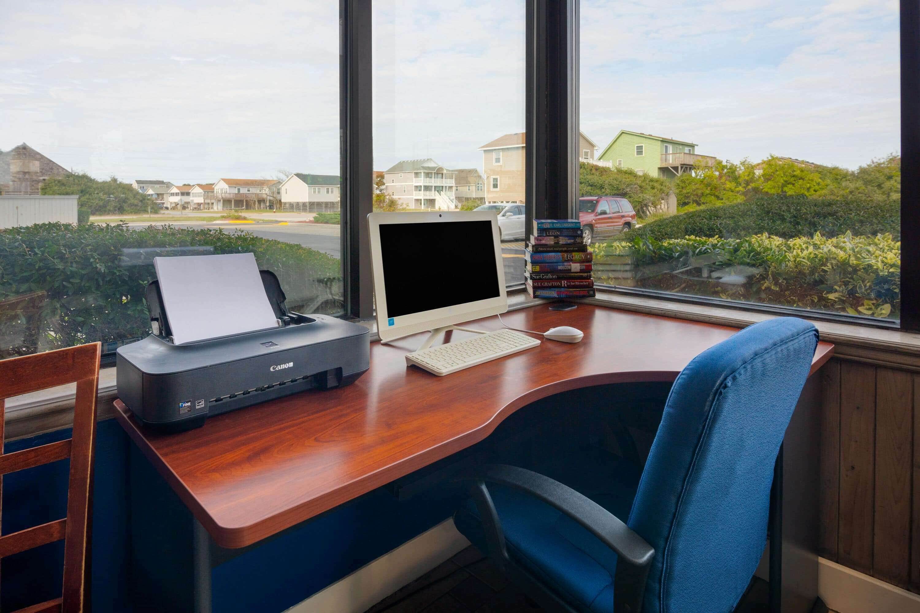 A wooden desk with a Canon printer, a white keyboard, a white mouse, and a monitor. There is a stack of books on the monitor's stand.