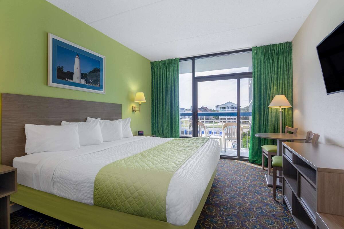 Habitación de hotel con una cama grande, un escritorio, una silla, un televisor, un balcón con vistas y un cuadro en la pared.