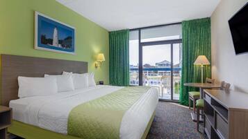 A hotel room with a large bed, a desk, a chair, a TV, a balcony with a view, and a painting on the wall.