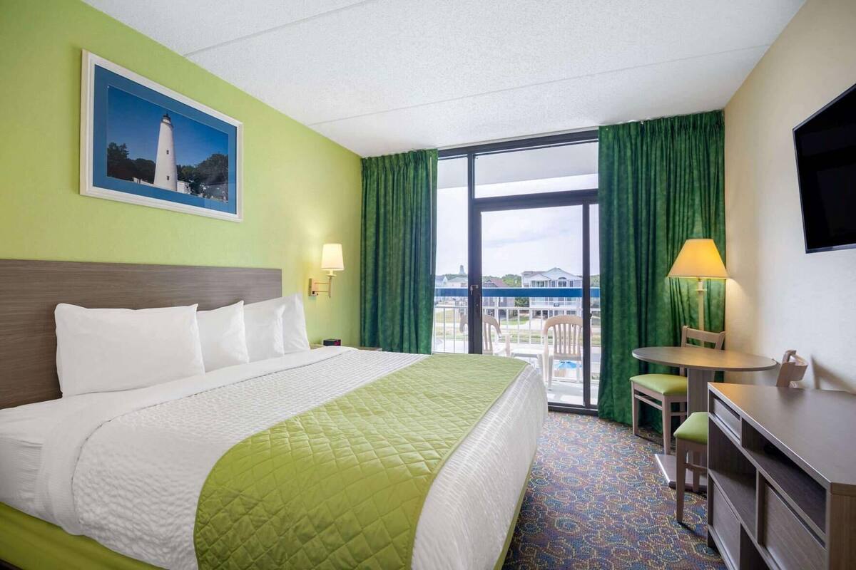 Habitación de hotel con cama, escritorio, silla, televisión, balcón con vistas y un cuadro en la pared.