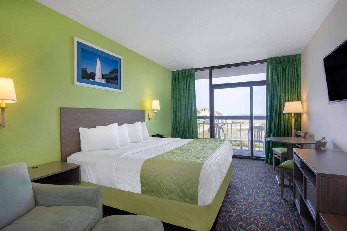 Habitación de hotel con cama, escritorio, silla, televisor y balcón con cortinas.