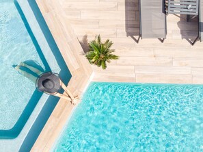 An aerial view of a swimming pool with clear blue water, a tiled deck, and a palm tree.