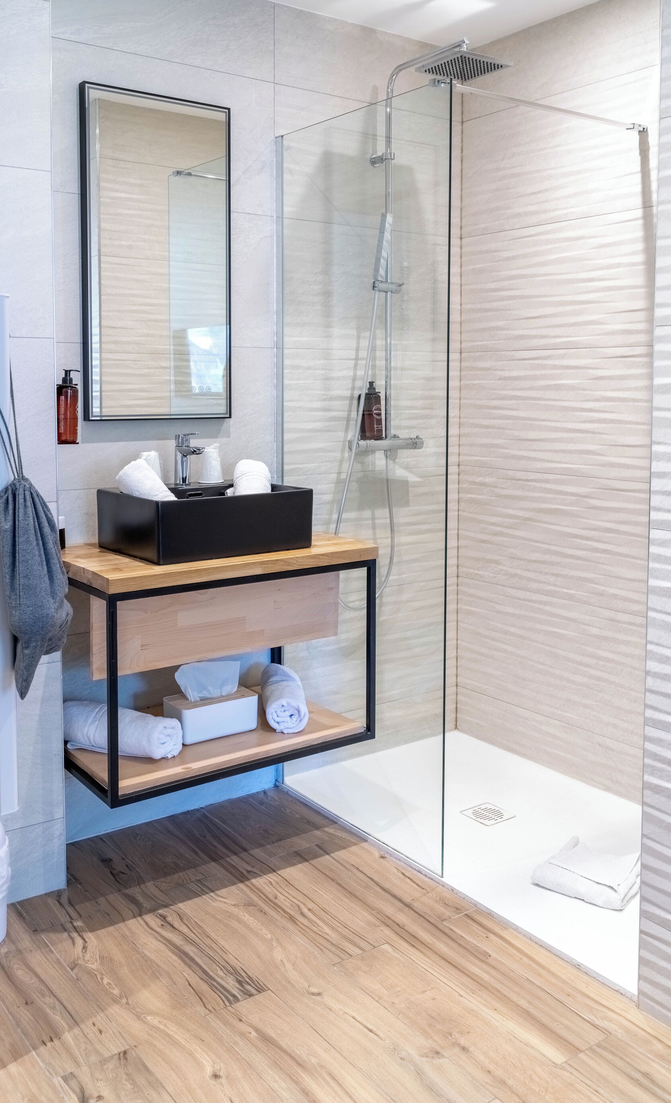 A modern bathroom with a glass shower enclosure, a wooden vanity with a black soap dispenser, and a mirror above the sink.
