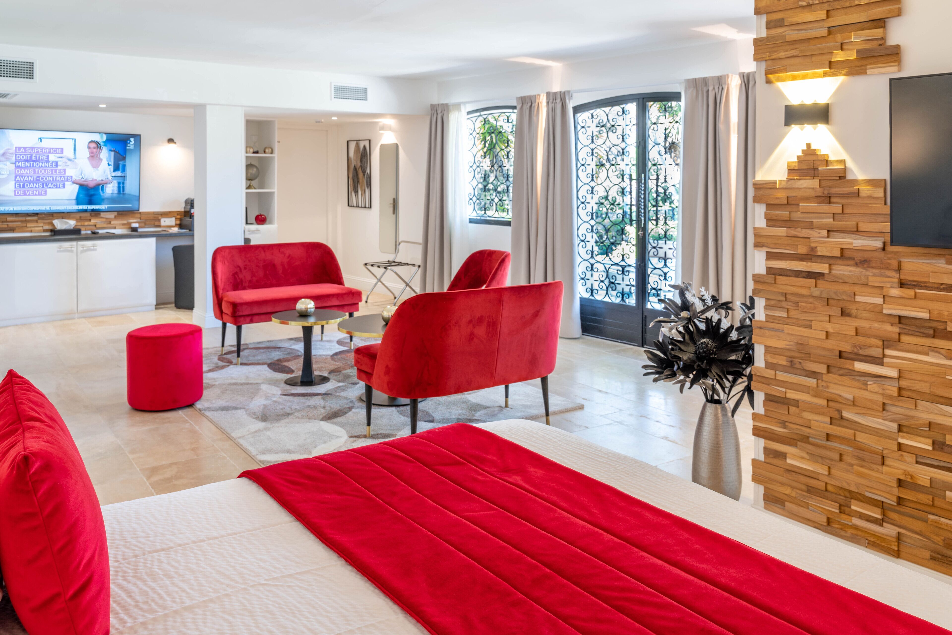 A modern hotel room with a large bed, red chairs, a TV, and a balcony with a view of greenery.