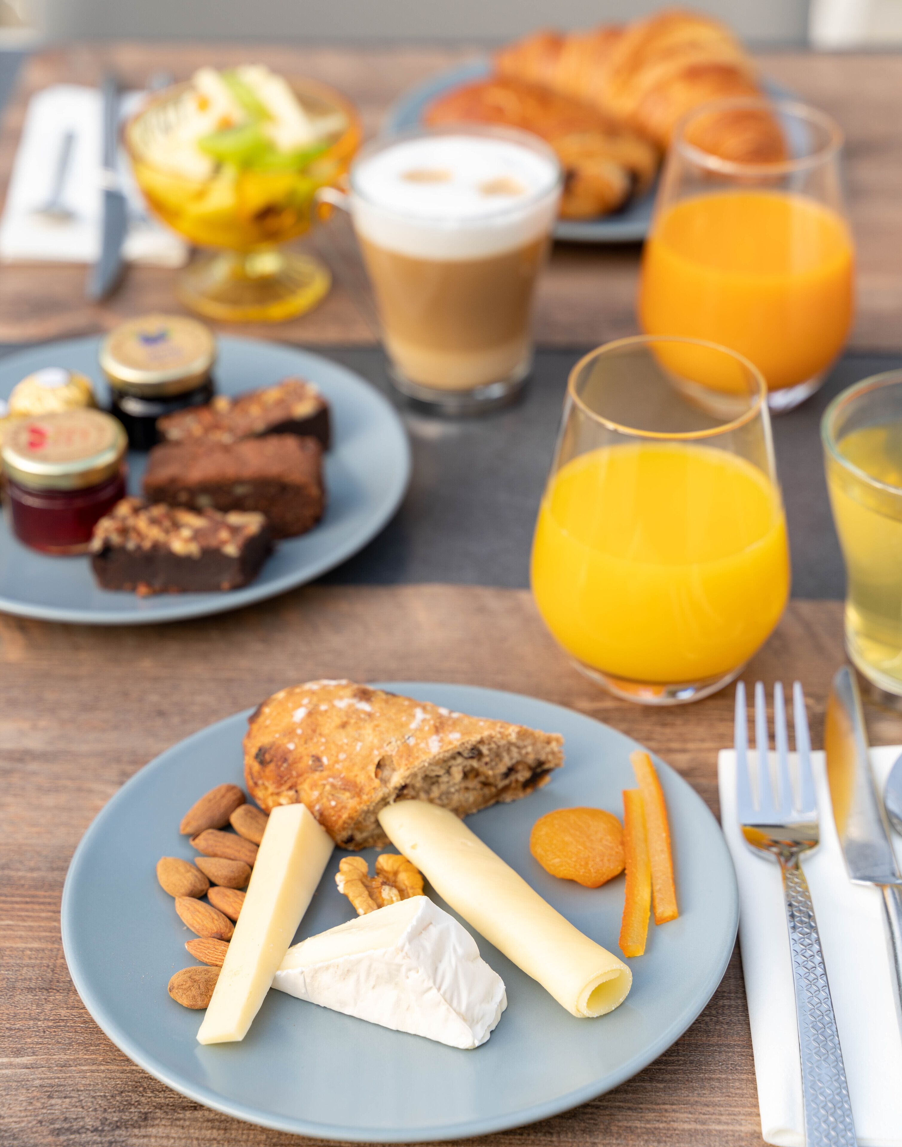 En frukostuppläggning med en tallrik mat som innehåller ost, nötter och ett bakverk, tillsammans med drycker och en croissant.