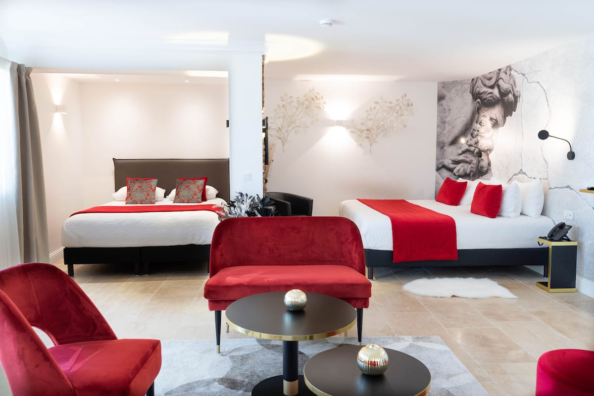 A modern hotel room with two beds, red and white bedding, a red sofa, and a round table with decorative balls.
