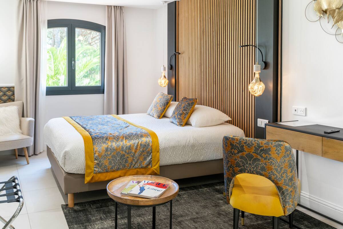A modern hotel room with a bed, a desk, two chairs, a round table, and a window with curtains.