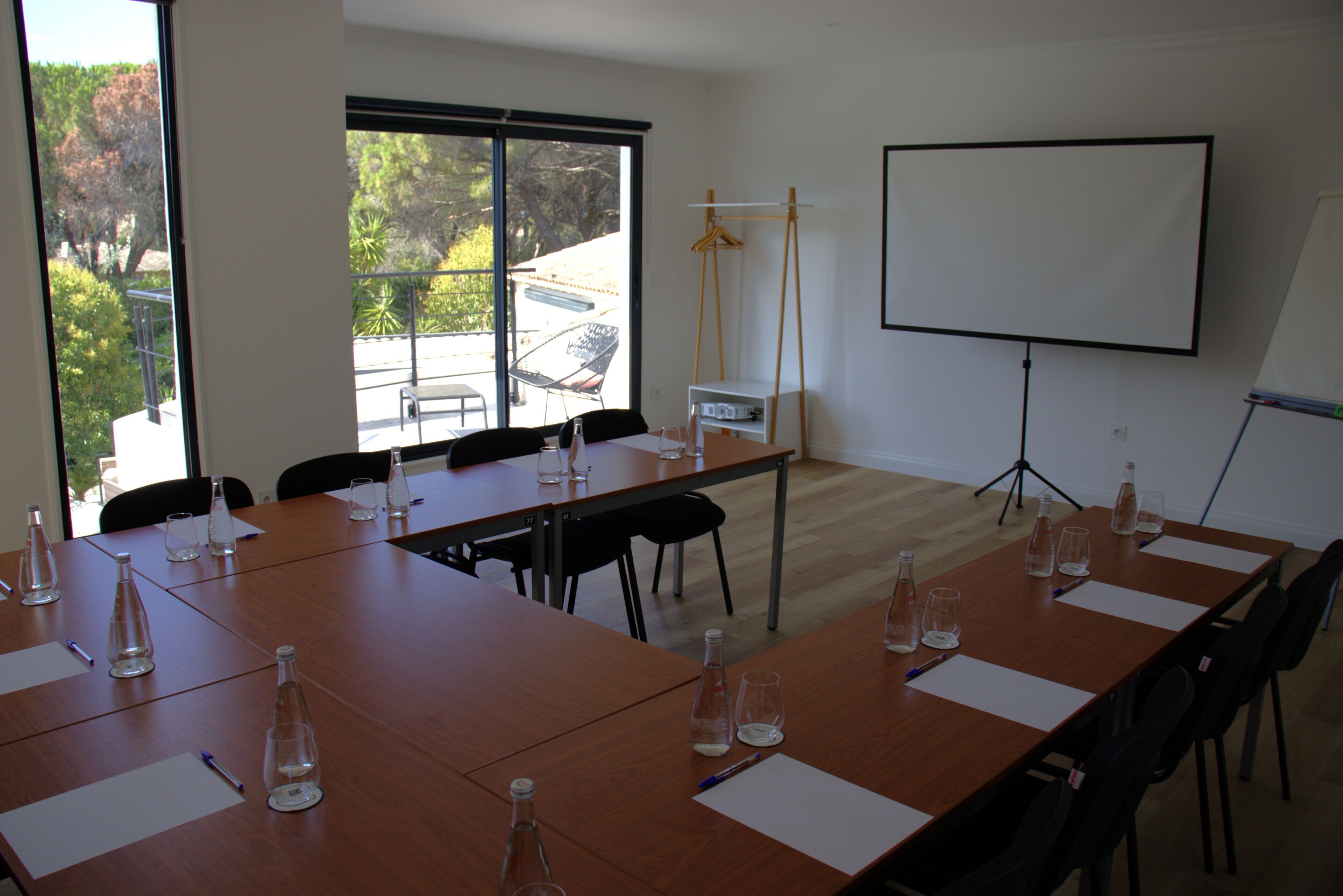 Un salón de conferencias con una larga mesa, sillas, una pantalla de proyector y una ventana con vistas a árboles y un edificio.