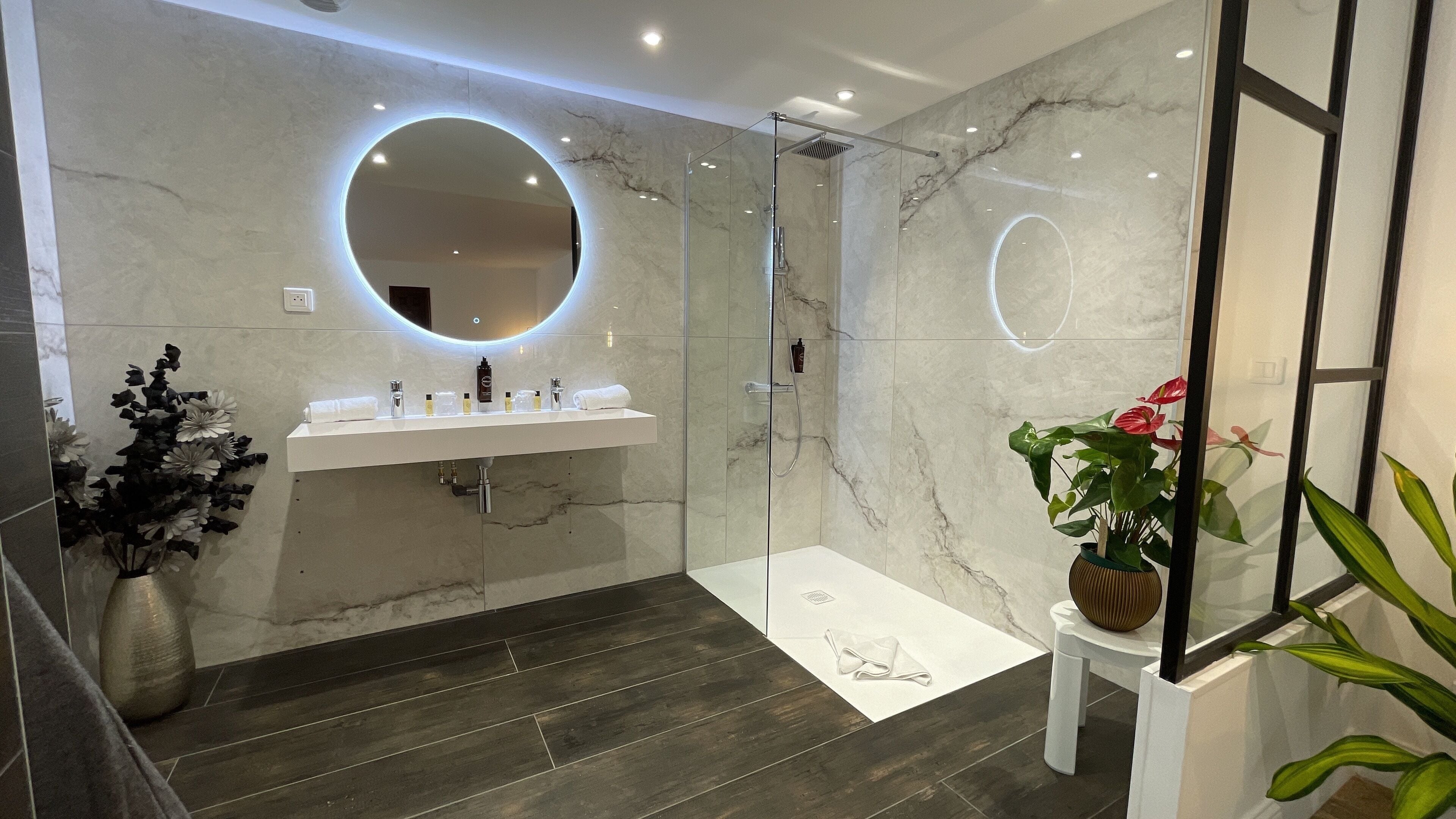 A modern bathroom with a glass-enclosed shower, a round mirror, a white sink with toiletries, and a dark wooden floor.
