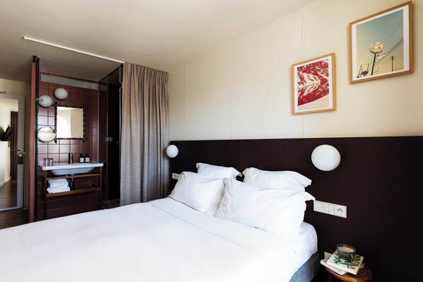 A hotel room with a large bed, a bedside table with a lamp, a mirror, a bathroom with a sink and towel rack, and two framed pictures on the wall.