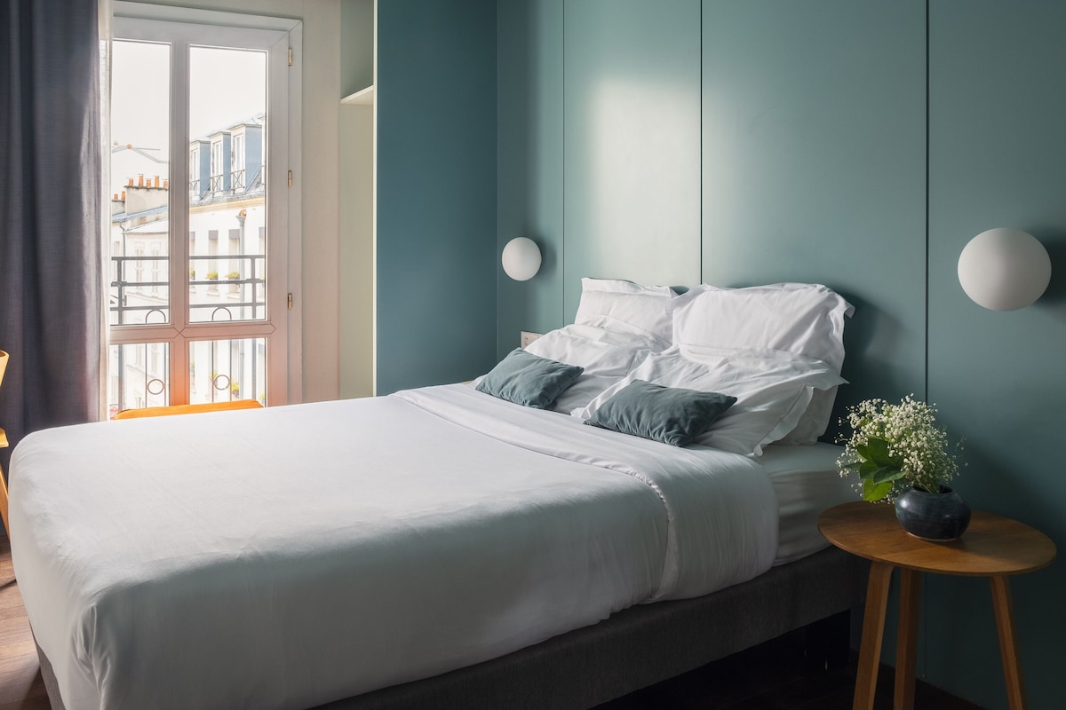 A bedroom with a bed, a chair, a small table, and a view of buildings through a window.