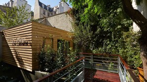 A wooden structure with a sign, a metal railing, and a red floor, surrounded by greenery and buildings in the background. - Hotel Cabane - Orso Hotels (Paris)