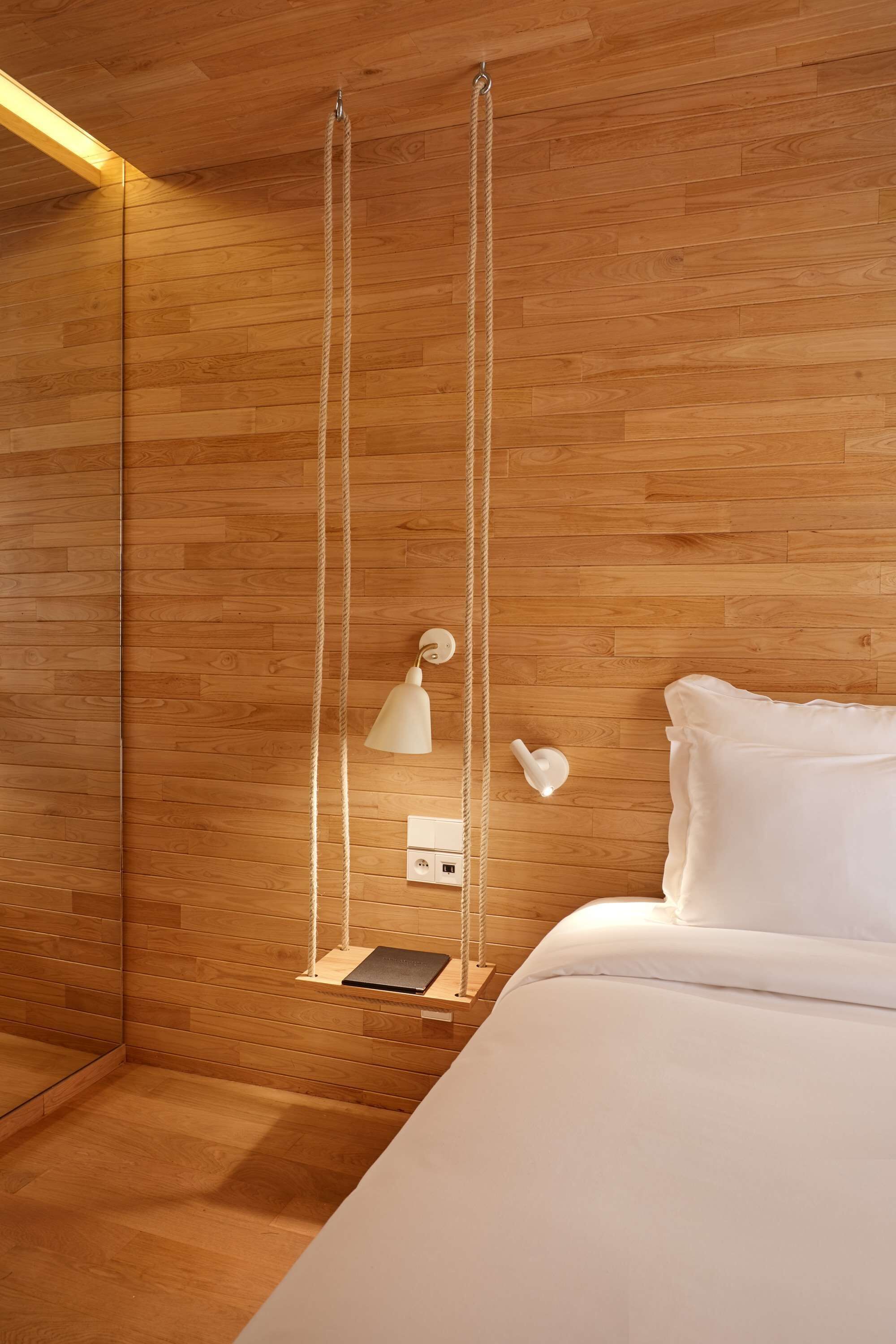 A bedroom with a wooden floor, a bed with white bedding, a white lamp, and a hanging swing with a book.