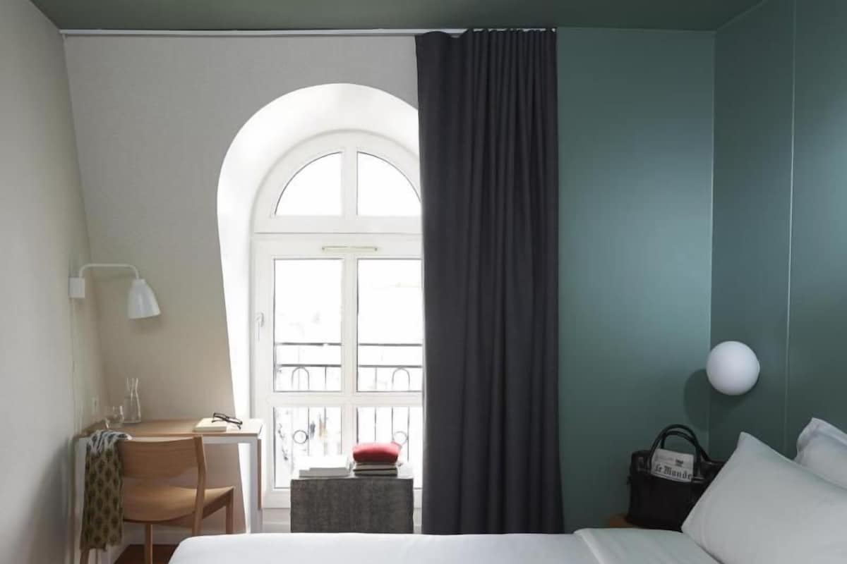 A hotel room with a bed, a desk with a chair, a small table, a window with curtains, and a view of the outside balcony.