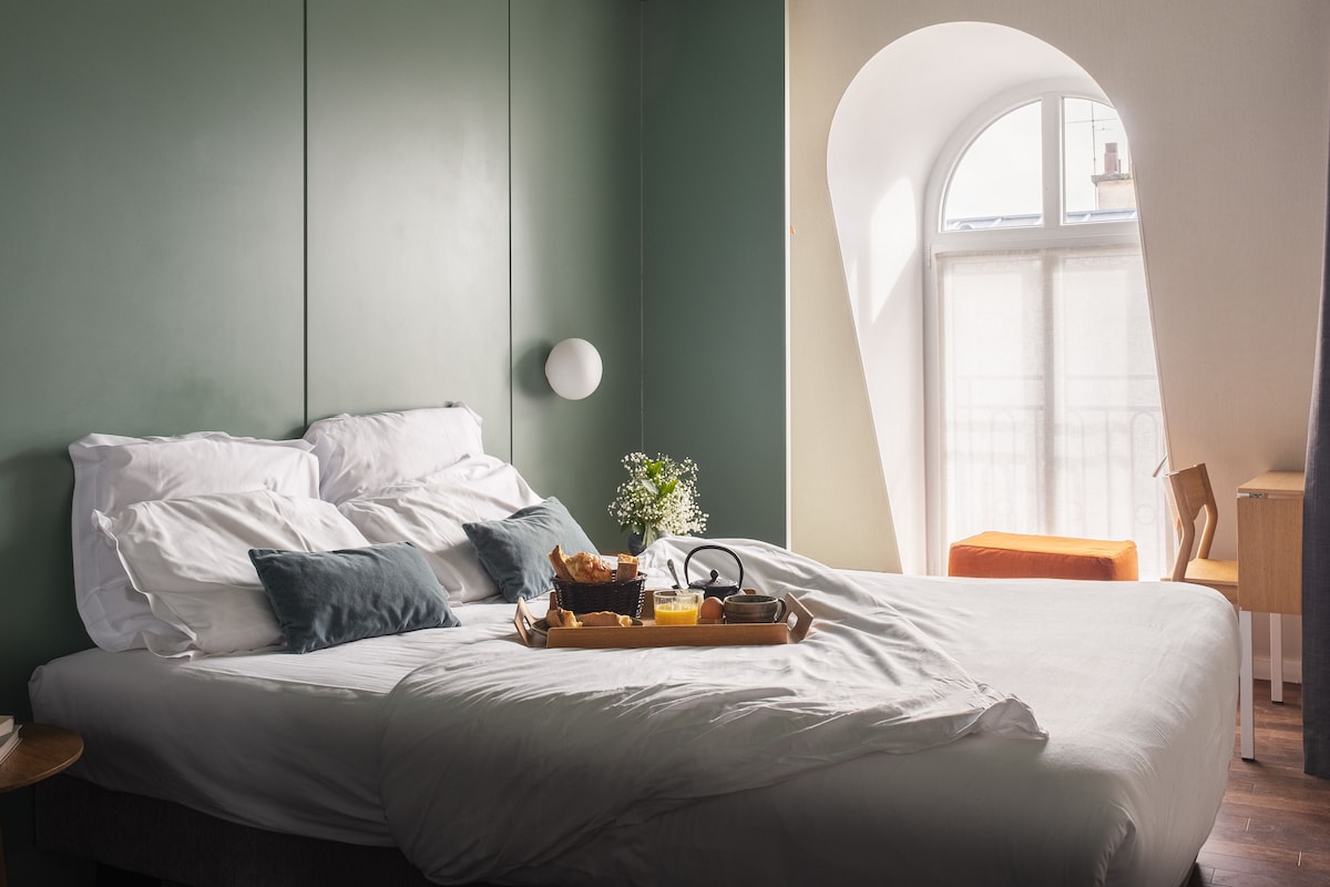 A neatly made bed with pillows, a tray with breakfast items, a vase with flowers, and a view of the city through a window.