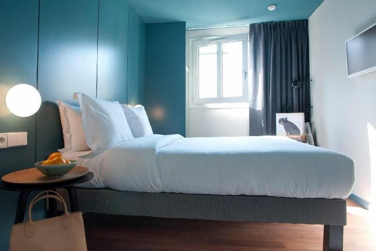 A bedroom with a bed, a bedside table with a bowl of fruit, a white bag, and a window with curtains.