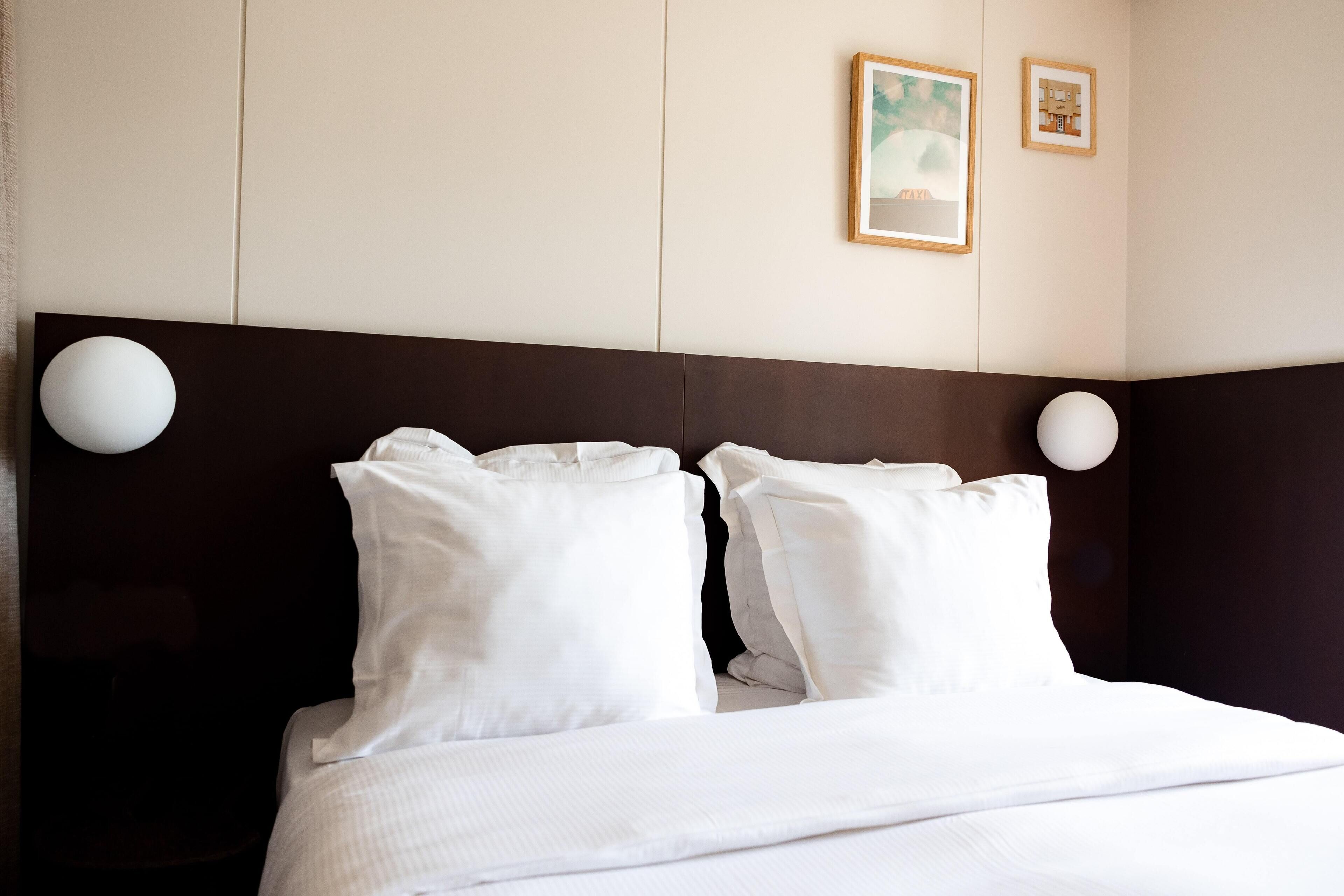 A hotel room with a large bed, two white pillows, a dark headboard, and two wall-mounted light fixtures.