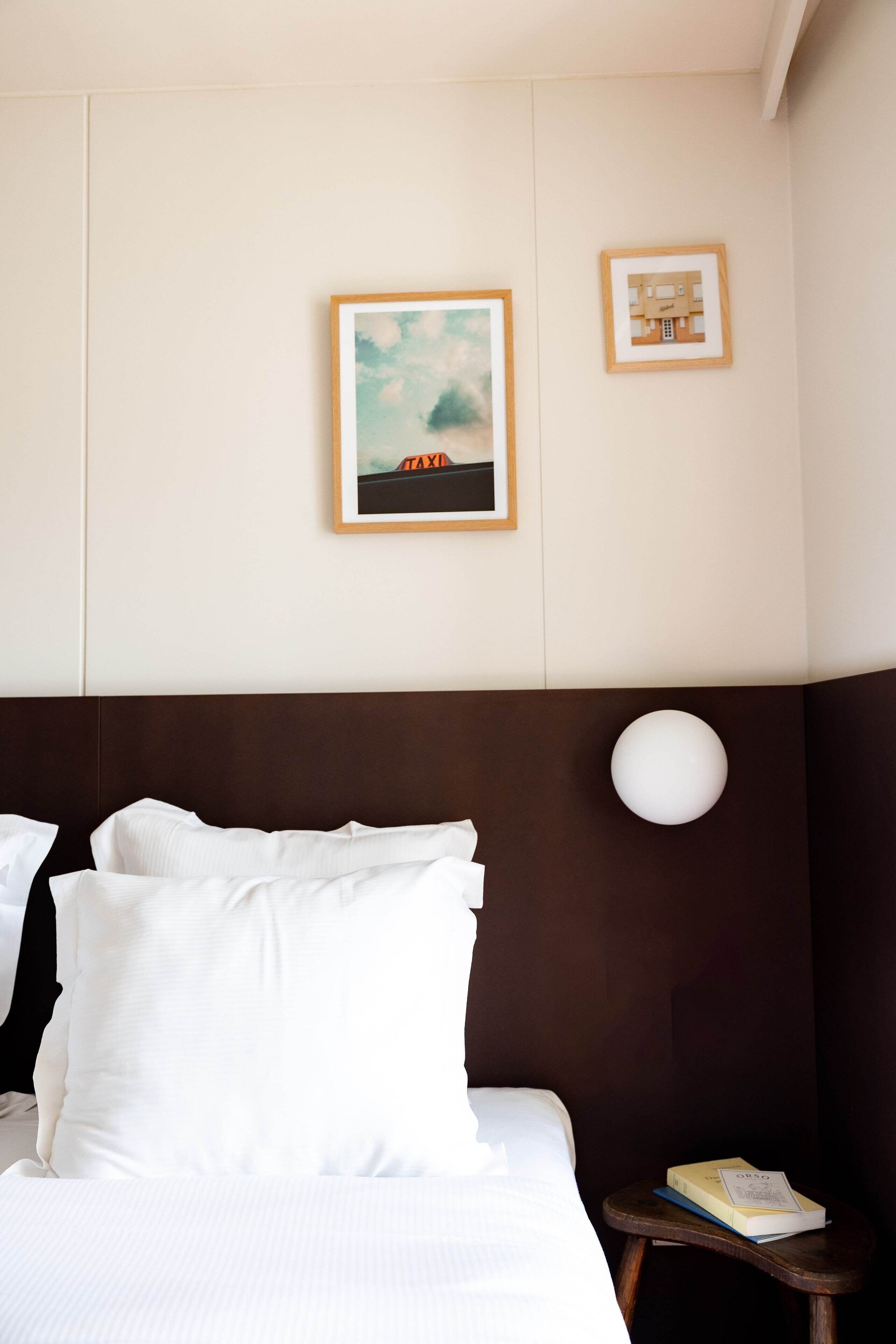 A hotel room with a bed, pillows, a nightstand with a book, a wall-mounted lamp, and two framed pictures.