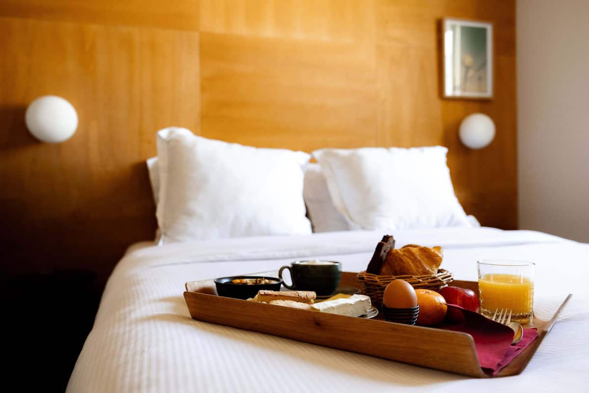 Una cama bien tendida con sábanas blancas, cabecera de madera y una bandeja de desayuno con cruasanes, huevos, fruta y zumo de naranja en una habitación de hotel.