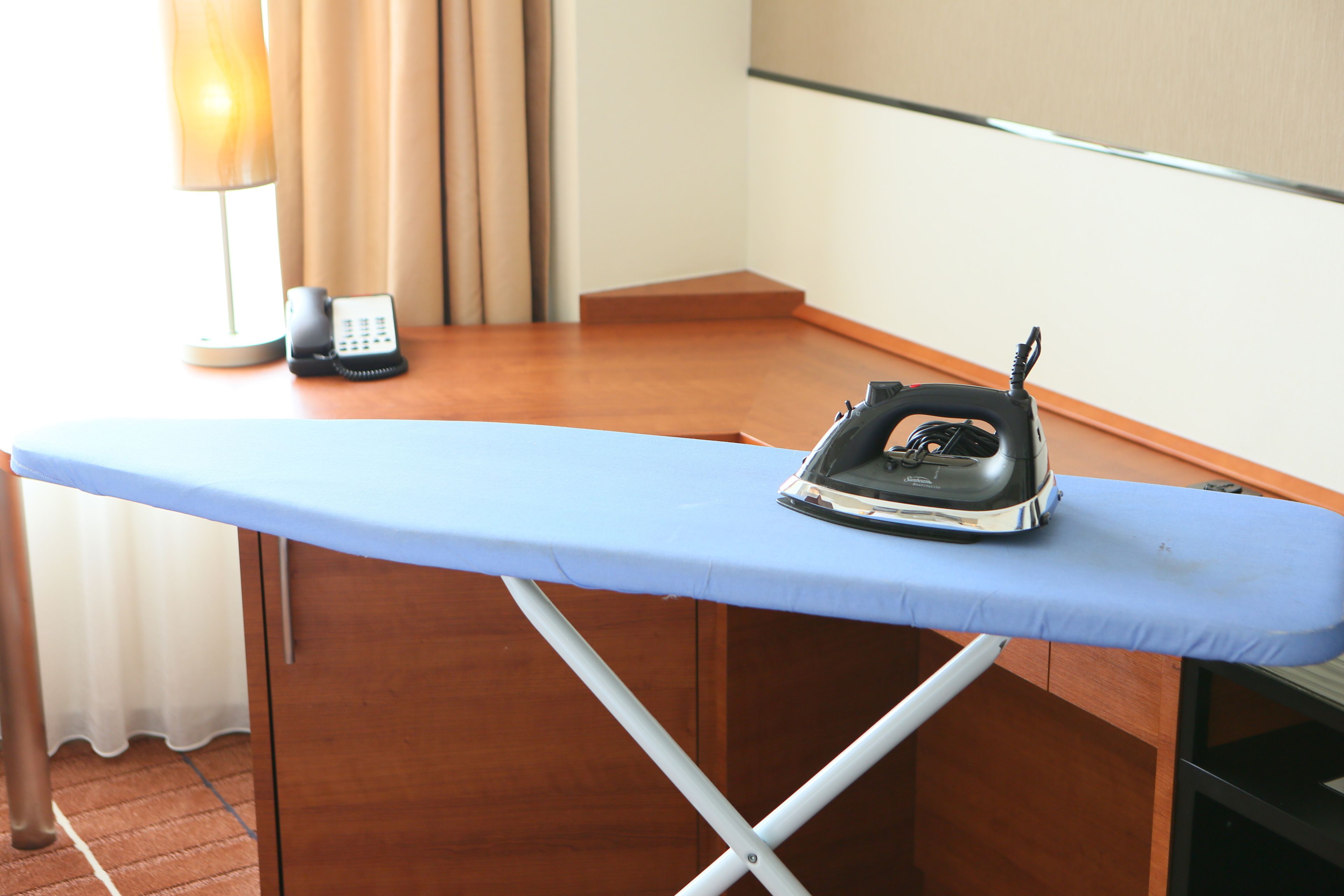 A room with a wooden desk, an ironing board with an iron, a telephone, and a lamp.