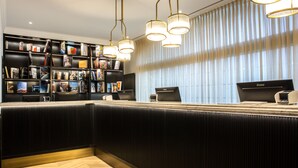Un moderno hall de recepción de hotel con un mostrador negro, una estantería llena de libros y revistas, y un ventanal con persianas verticales.
