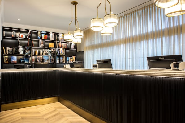 A modern hotel reception area with a black reception desk, a bookshelf filled with books and magazines, and a large window with vertical blinds. - a&o Brussel Centrum   (Brussels)
