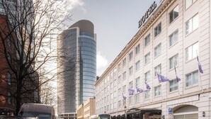 Un hotel con fachada clásica, un edificio moderno de vidrio al fondo y un autobús verde estacionado al frente.