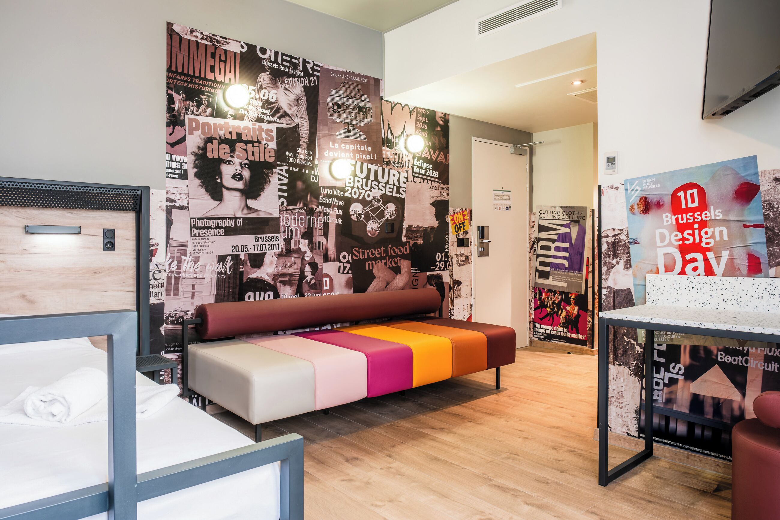 A modern hotel room with a colorful bench, a bed with a wooden headboard, and a wall decorated with various posters.