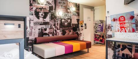 A modern hotel room with a colorful bench, a bed with a wooden headboard, and a wall decorated with various posters.