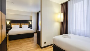Habitación de hotel con dos camas, suelo de madera, cabecera moderna, lámpara de noche y ventana con cortinas.