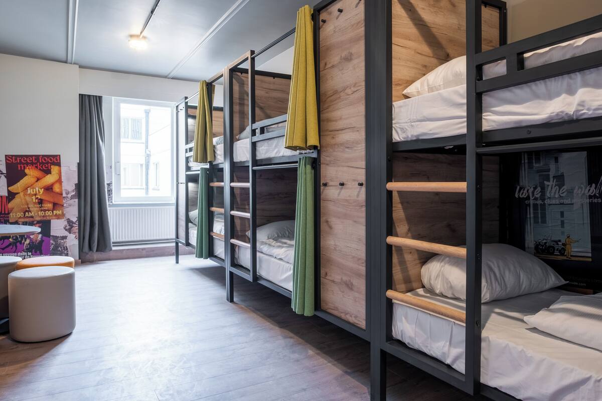A hostel room with bunk beds, a window with curtains, a poster on the wall, and a small stool.