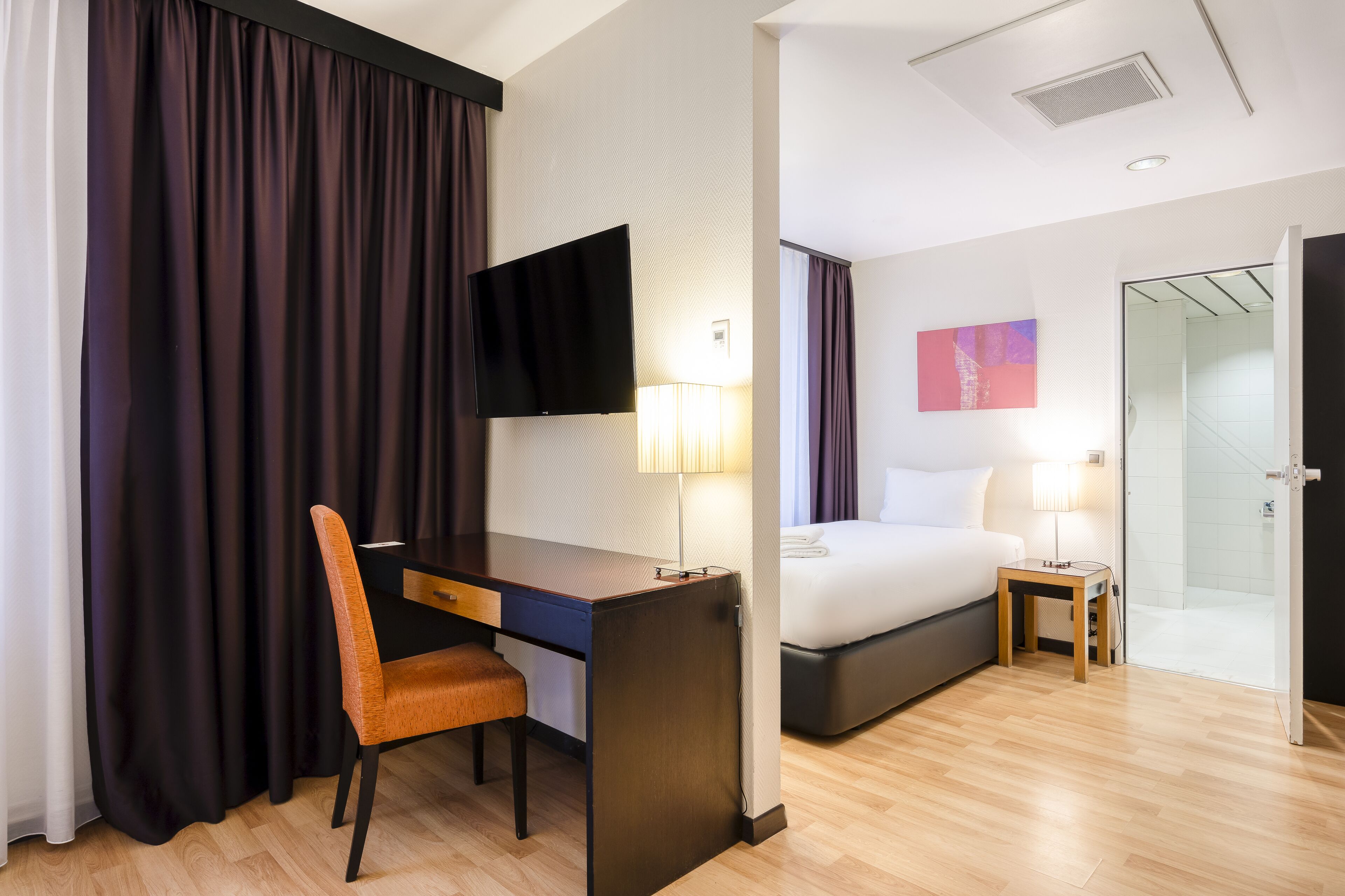 A hotel room with a desk, chair, bed, bedside table, lamp, TV, and a shower area.