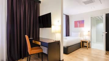 A hotel room with a desk, chair, bed, bedside table, lamp, TV, and a shower area.