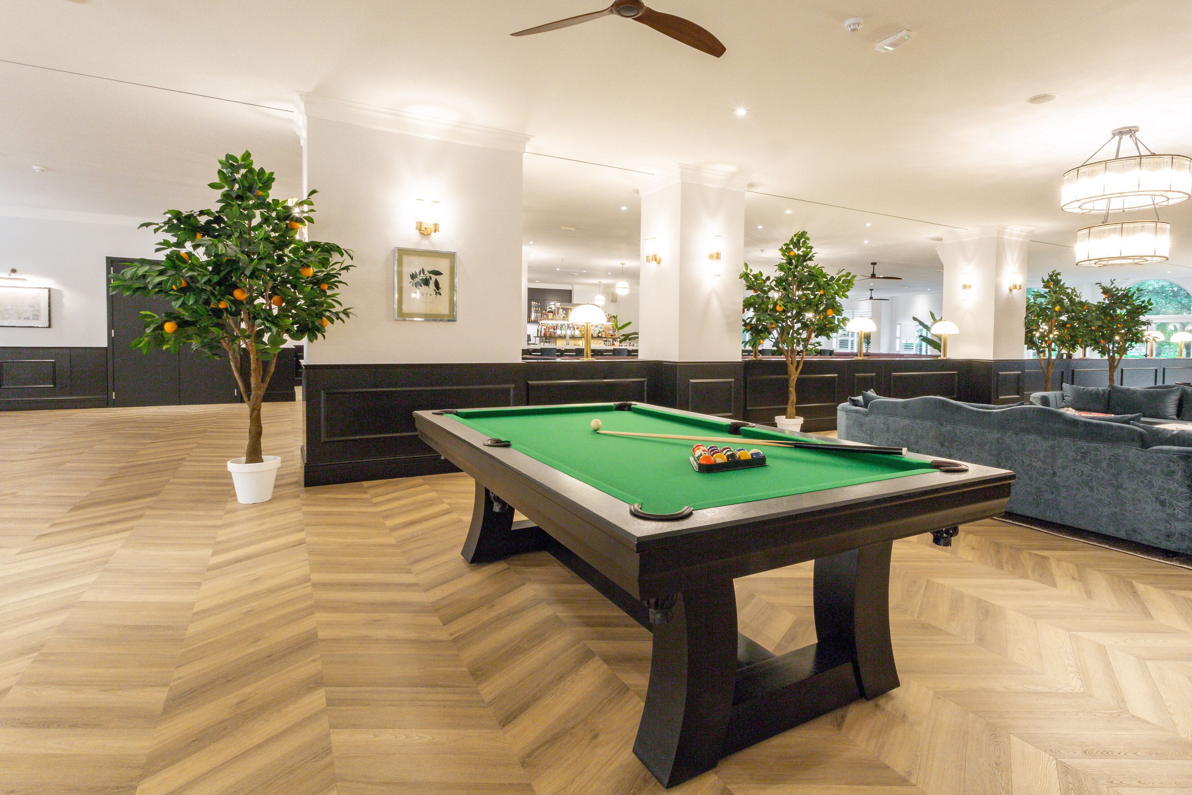 Une table de billard dans une pièce bien éclairée, dotée d’un parquet à motifs en chevrons, entourée de sièges et de plantes en pot.