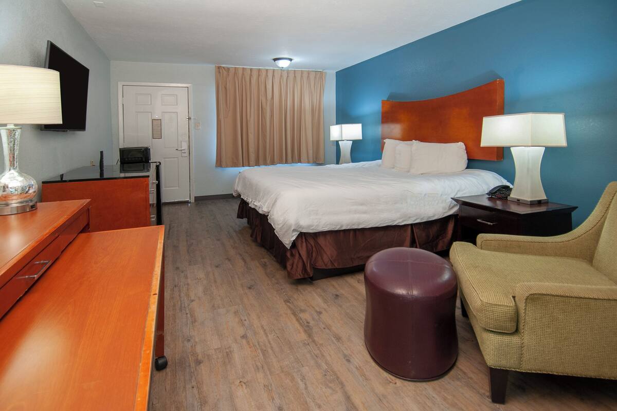 A hotel room with a bed, two bedside lamps, a desk with a lamp, a chair, and a small ottoman.
