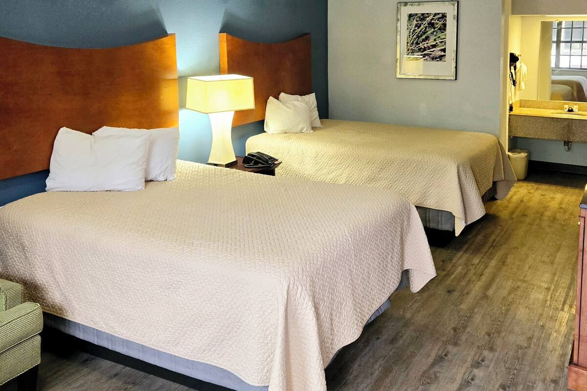 A hotel room with two beds, a wooden floor, a headboard with a light fixture, a bedside table with a lamp, and a framed picture on the wall.