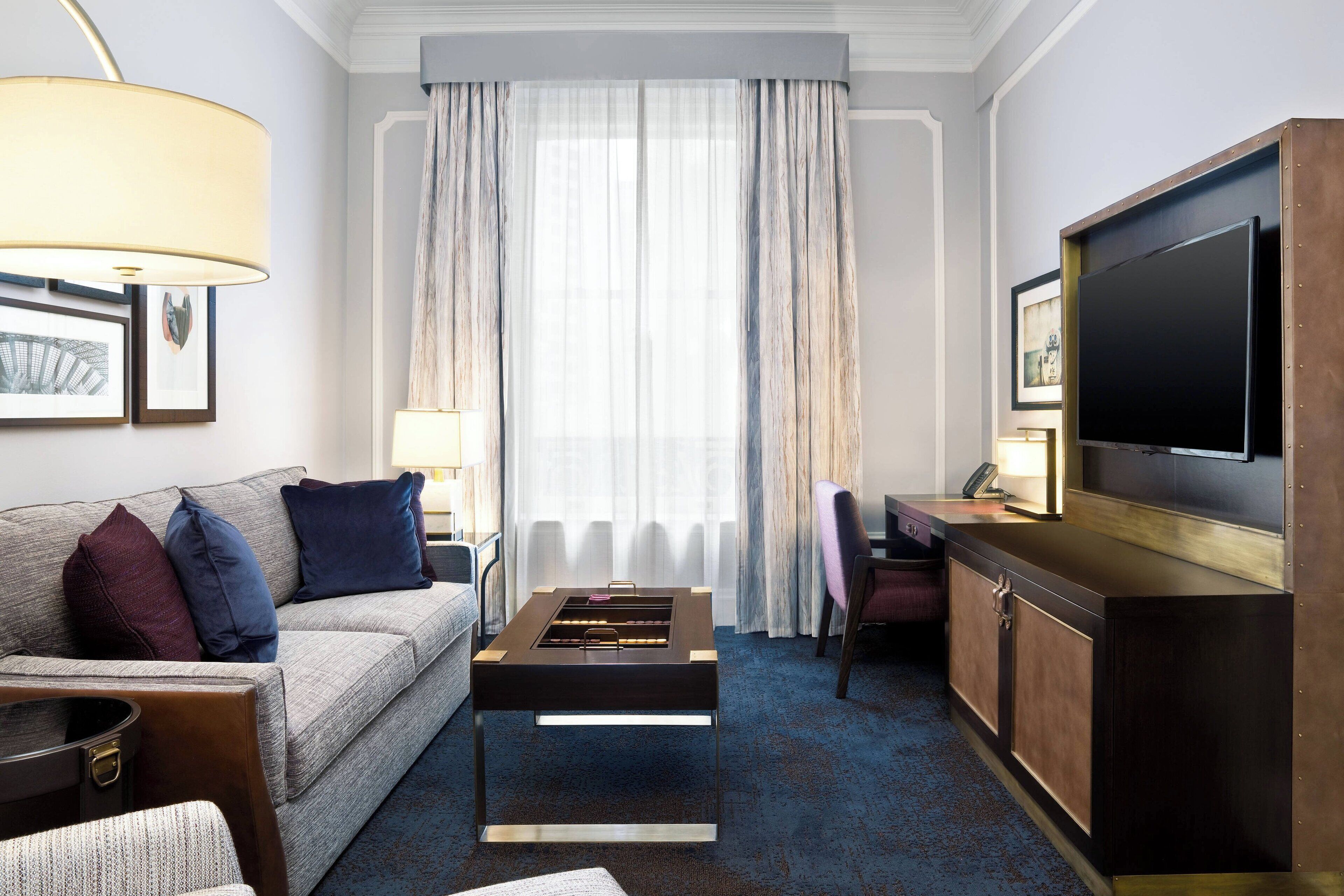 A hotel room with a sofa, a coffee table, a TV, a desk, and a large window with curtains.