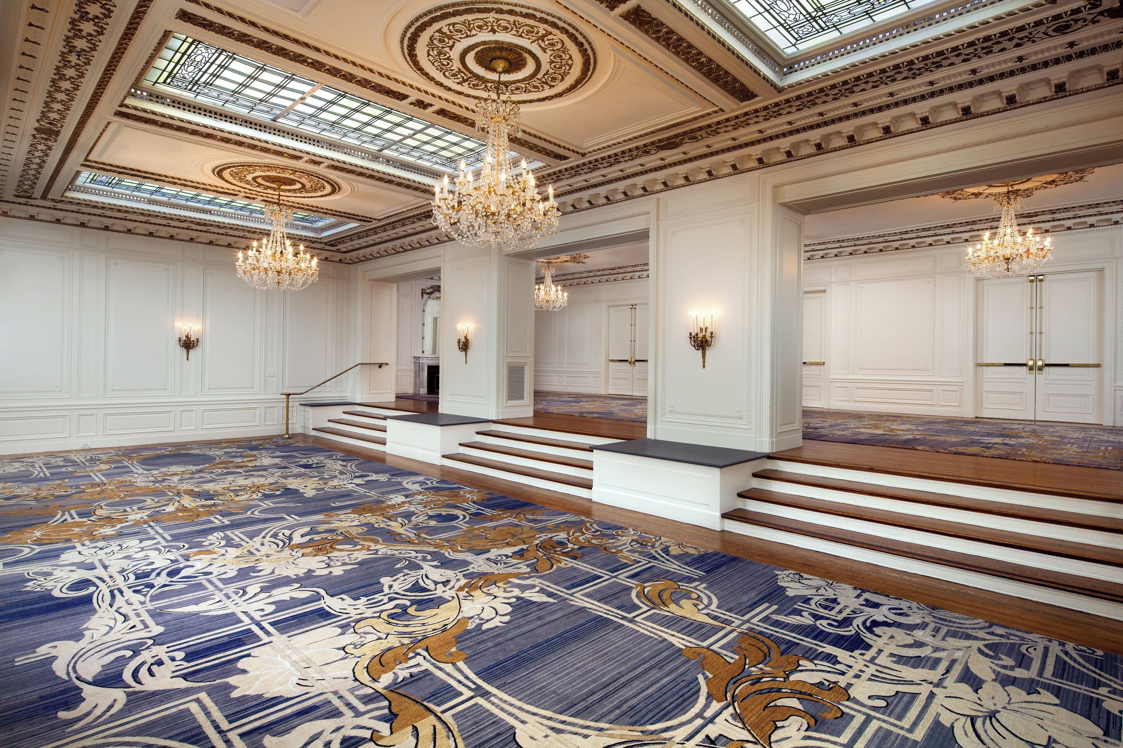 A grand hall with ornate chandeliers, a patterned carpet, and a staircase leading to a balcony.