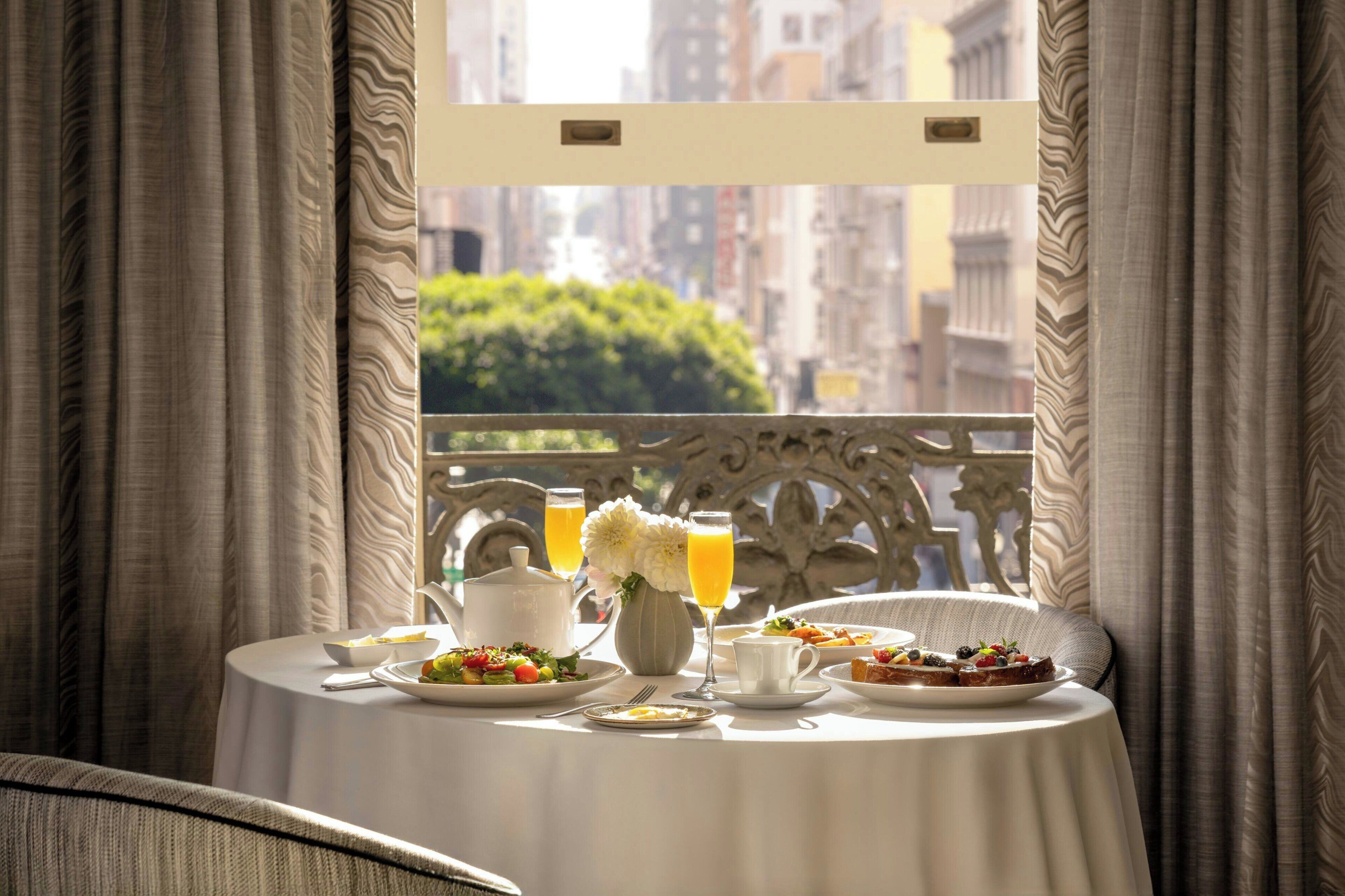Une table à manger avec des aliments pour le petit-déjeuner, un vase rempli de fleurs et une vue sur le paysage urbain à travers une fenêtre ornée de rideaux.