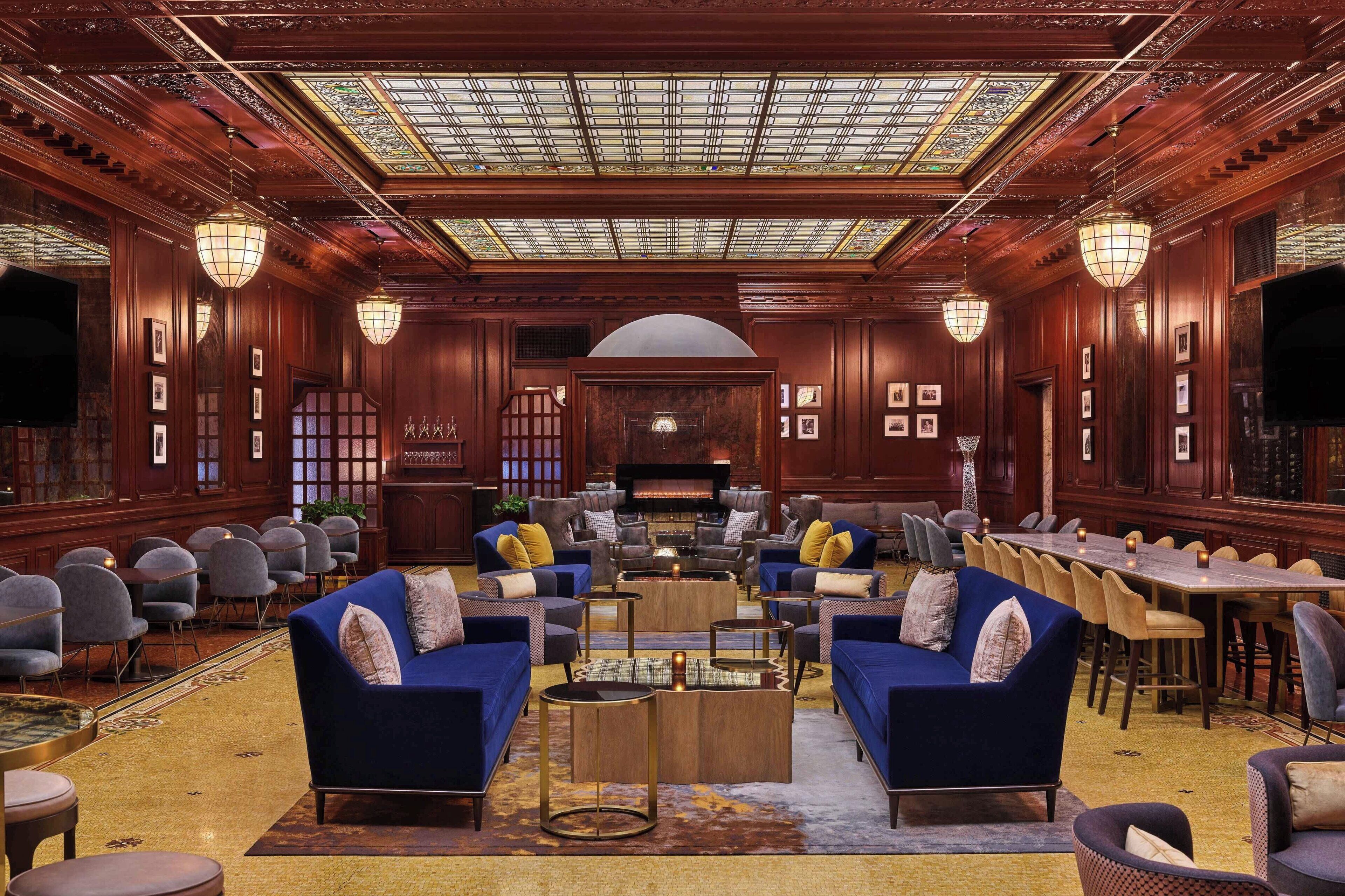 A spacious lobby with a high ceiling, ornate wood paneling, and a central fireplace. There are multiple seating areas with sofas and chairs, and a long dining table.