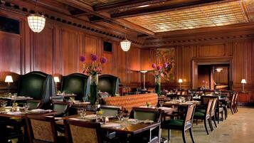 A formal dining room with wooden paneling, green upholstered chairs, and tables set for service.
