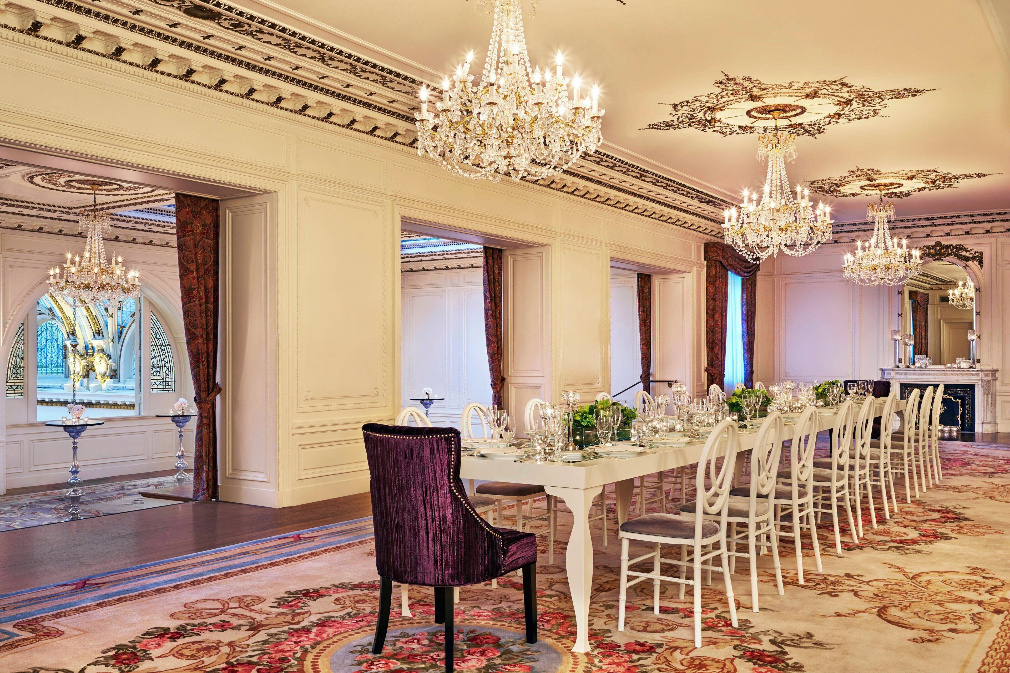 A formal dining room with a long table set for a meal, ornate chandeliers, and a richly patterned carpet.