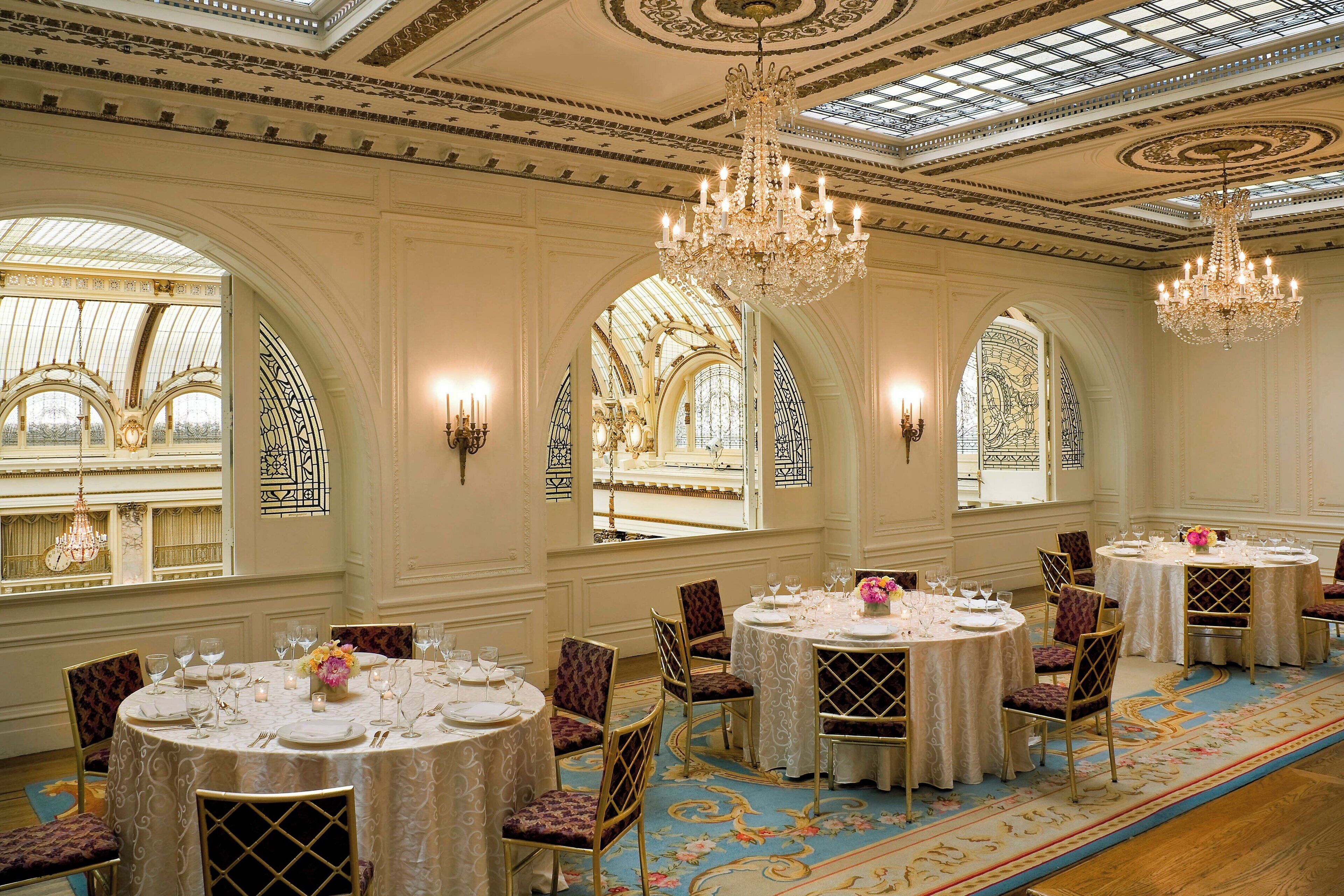 Un amplio comedor con candelabros ornamentados, mesas redondas dispuestas para comer y grandes ventanales con diseños intrincados.