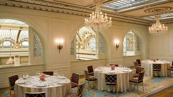 Un amplio comedor con candelabros ornamentados, mesas redondas dispuestas para comer y grandes ventanales con diseños intrincados.