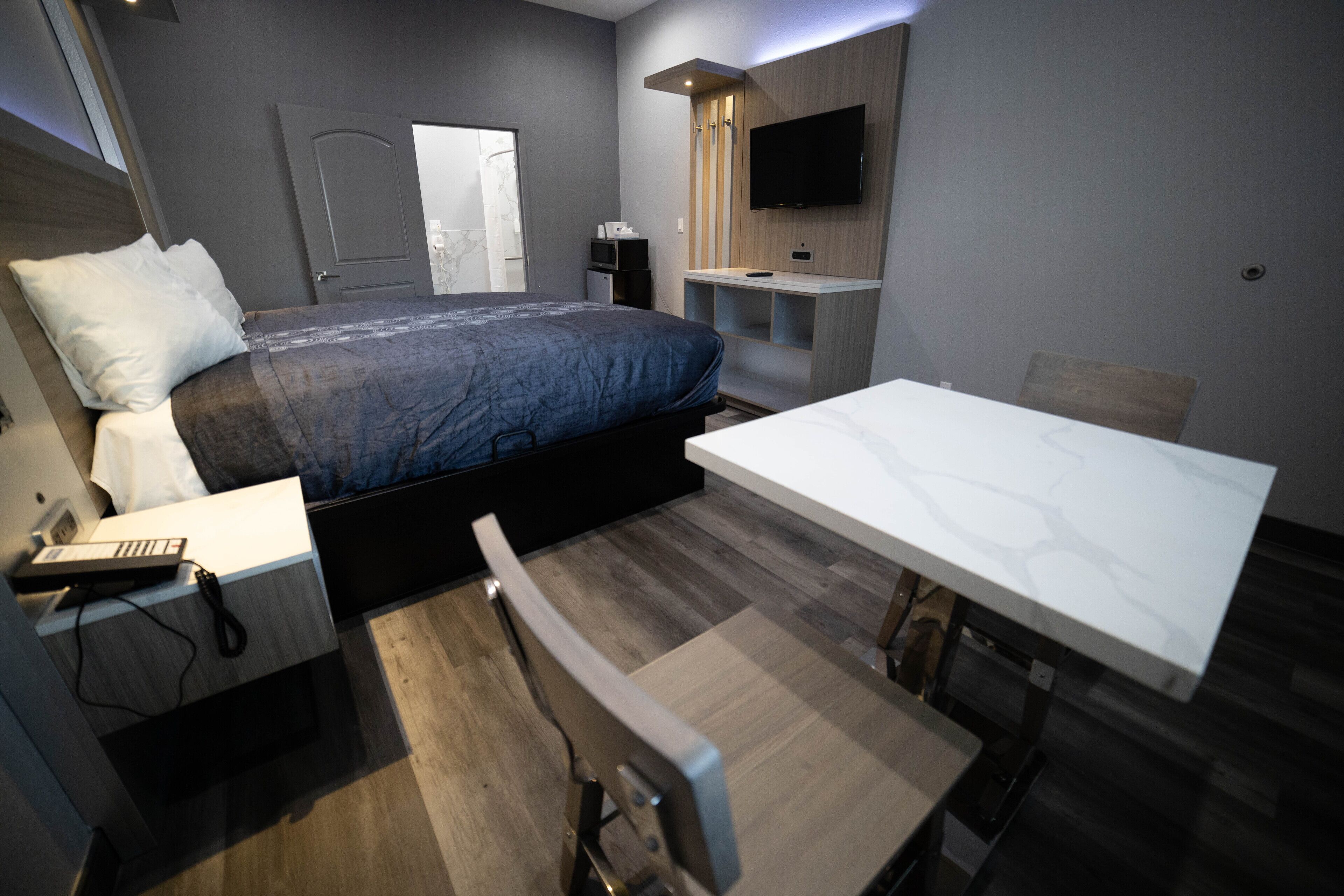 A modern hotel room with a bed, bedside table, desk, chair, and a TV mounted on the wall.