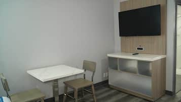 A small room with a white table, a wooden chair, a TV mounted on the wall, and a wooden cabinet with shelves.